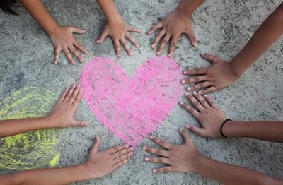 Children's hands surround a pink heart drawn in chalk on the sidewalk